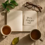 A soft, calming flat-lay of a journal, warm tea, and a note reading “You’re doing your best,” symbolizing mindfulness and emotional care.